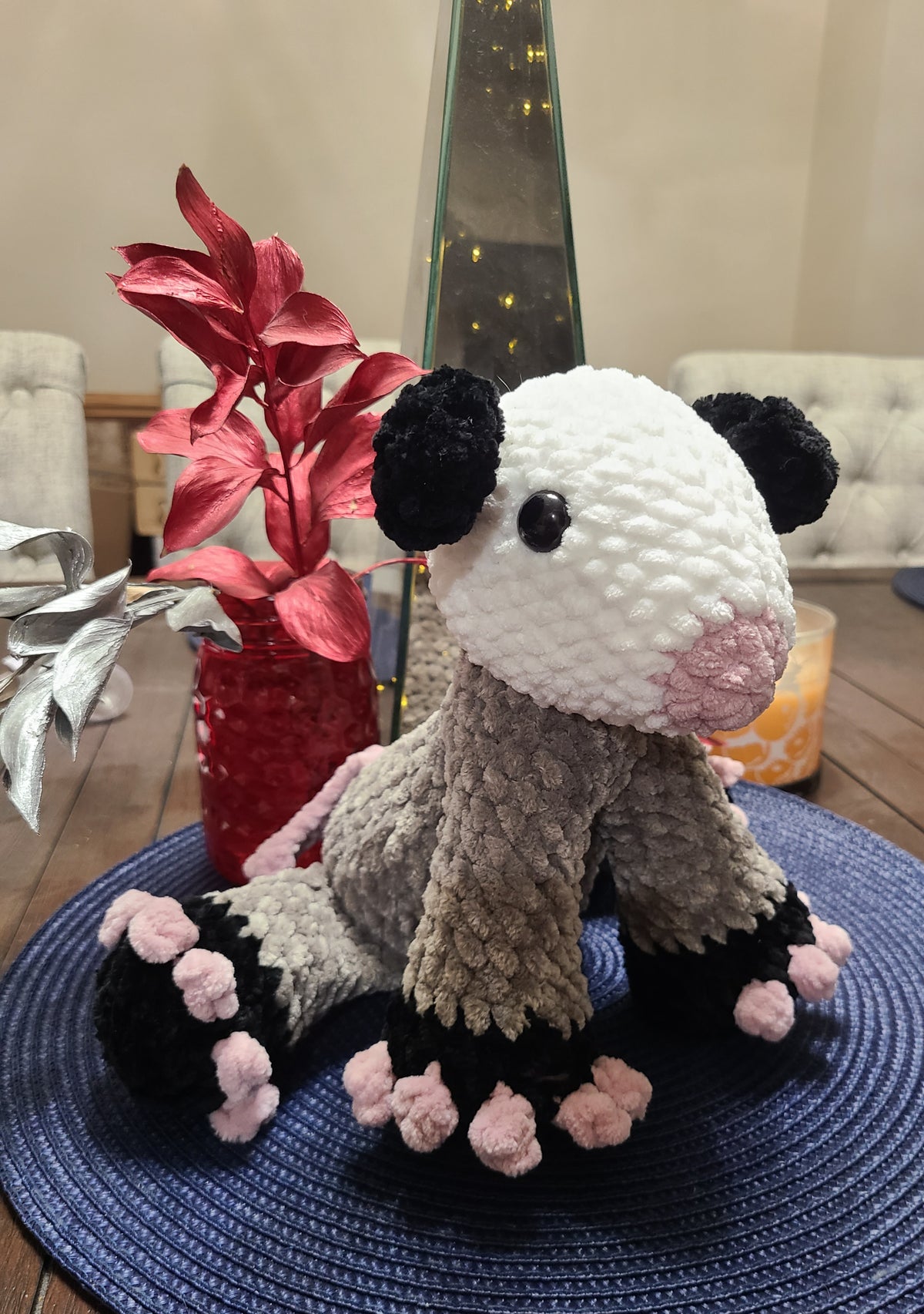 Crocheted plush opossum sitting on a blue textured surface with decorative elements in the background.