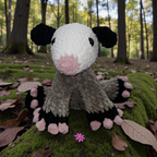 Crocheted plush opossum on a mossy forest floor with trees in the background