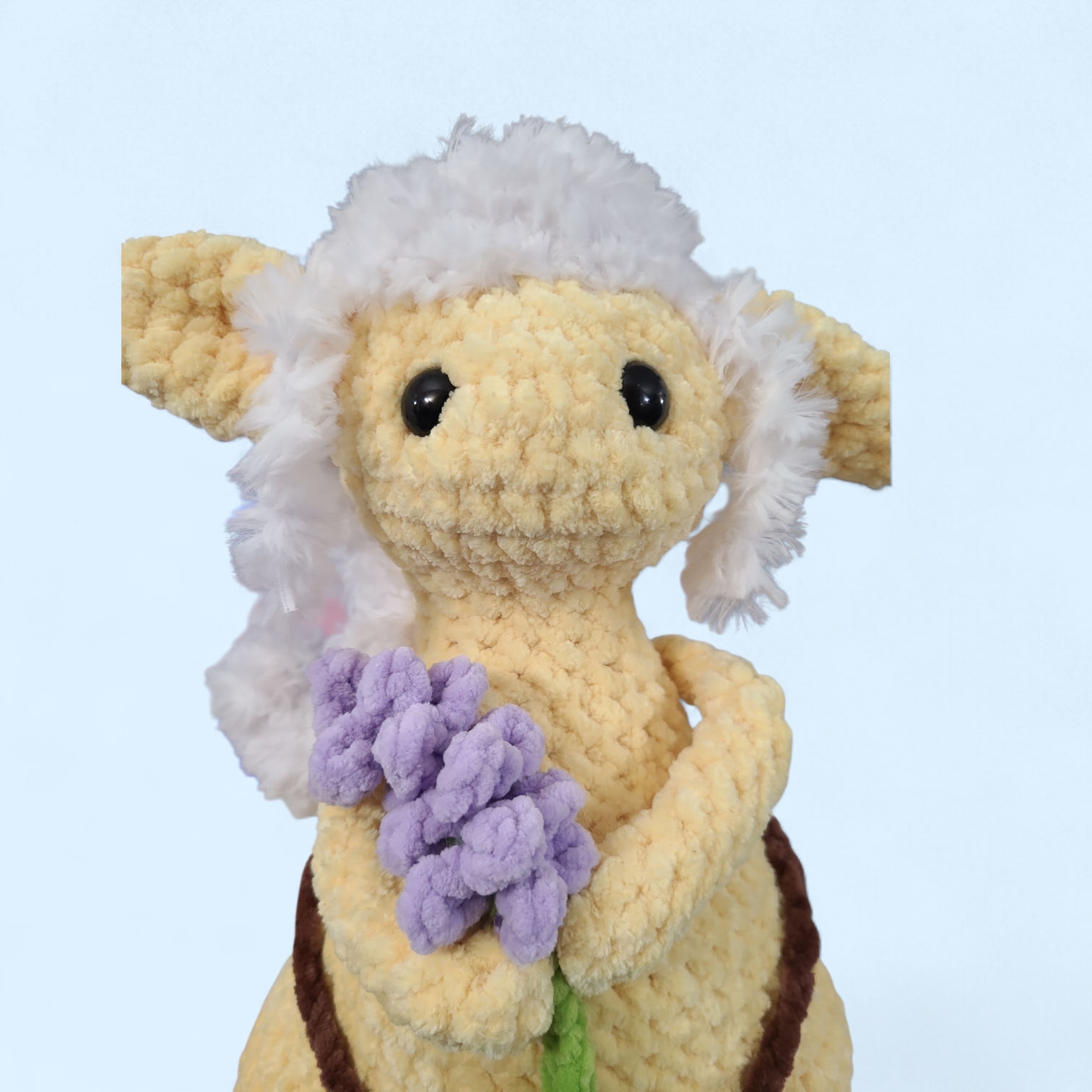 Crocheted yellow Forager creature  with white fur, holding a bundle of lavender on a light blue background