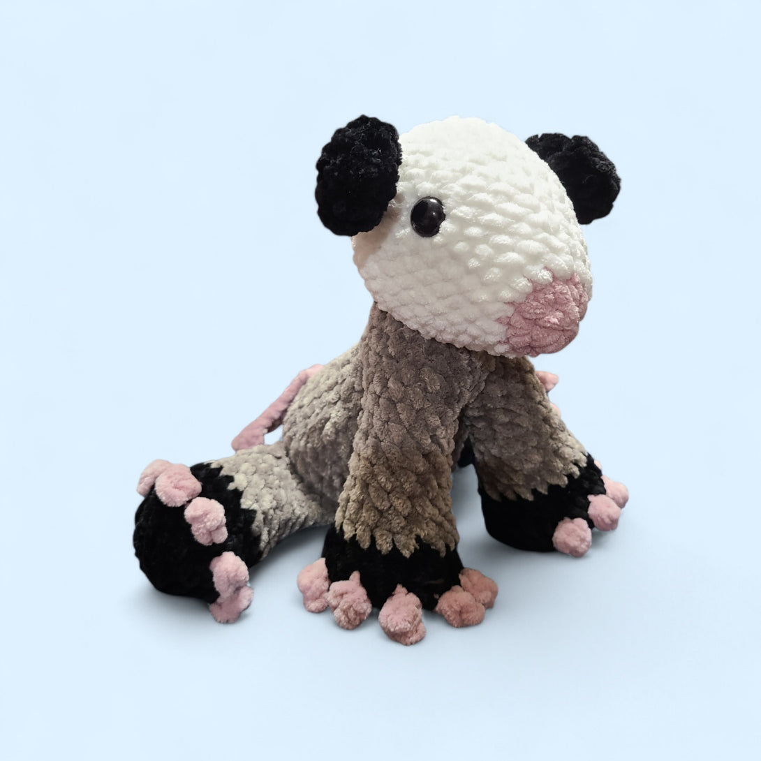 Crochet opossum in a seated position with a grey body, white head and pink toes on a light blue background
