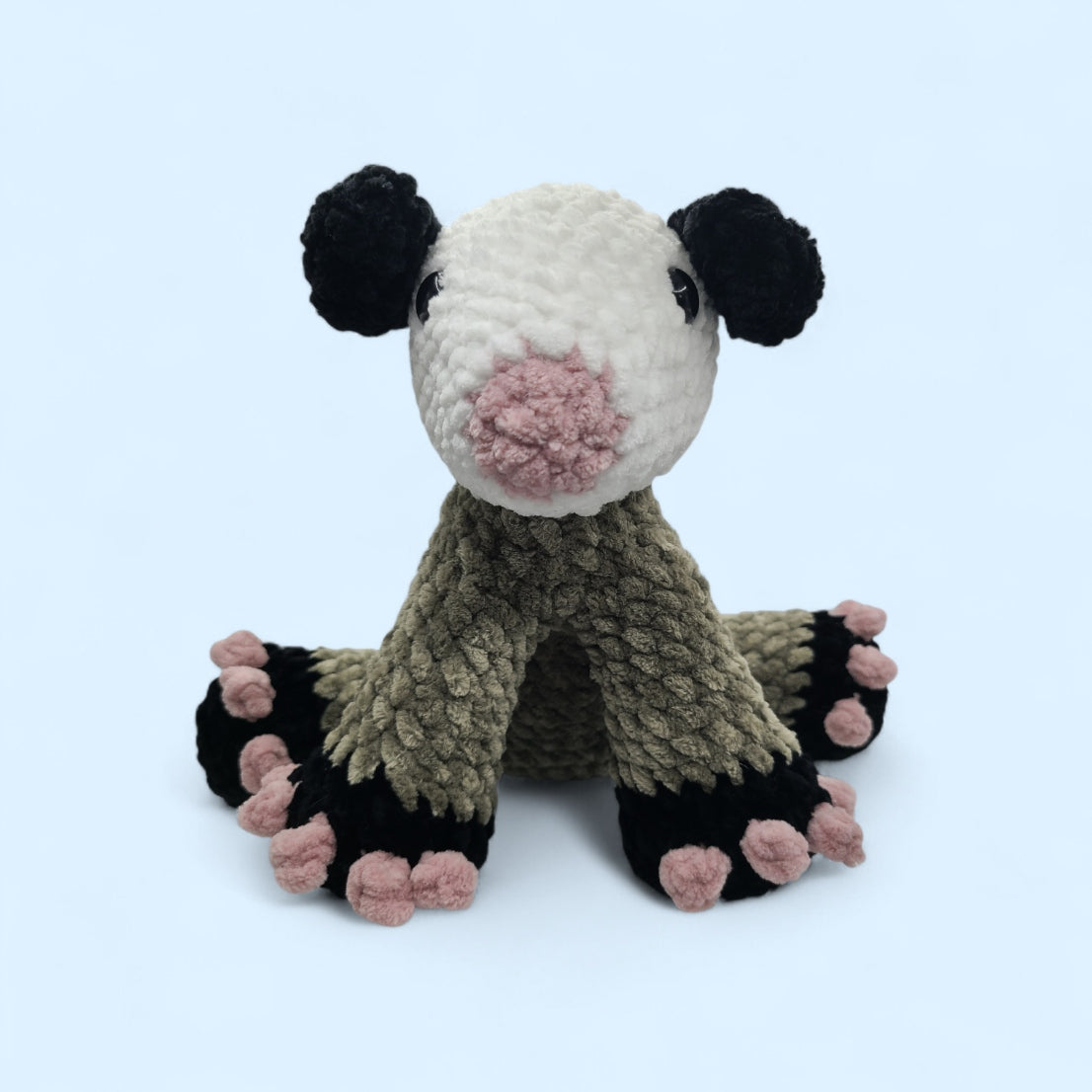 Crochet opossum in a seated position with a grey body, white head and pink toes on a light blue background